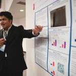 Confident young man in suit presenting research findings on a poster at a conference.