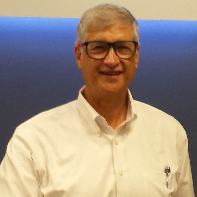 Smiling middle-aged man with gray hair and glasses in a white button-down shirt, standing against a blue gradient background, with a pen in his shirt pocket.