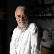 Older man in a white shirt leaning on a workbench in a workshop or lab setting behind him shelves and equipment visible at the back of the room.