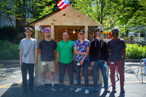 Six men standing in a row outside a wooden shed with a flag on top, smiling at the camera.