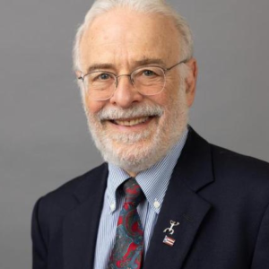 Smiling senior man with white hair and beard, glasses, wearing a navy blazer, striped shirt, patterned tie, and a lapel pin with the US flag.