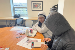 Two students in hoodies sit at a classroom table, working on a craft project with papers, rulers, and small tools on the desk.