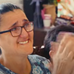 Smiling woman with glasses and dark hair in a bun, wearing a floral top, clapping beside her.