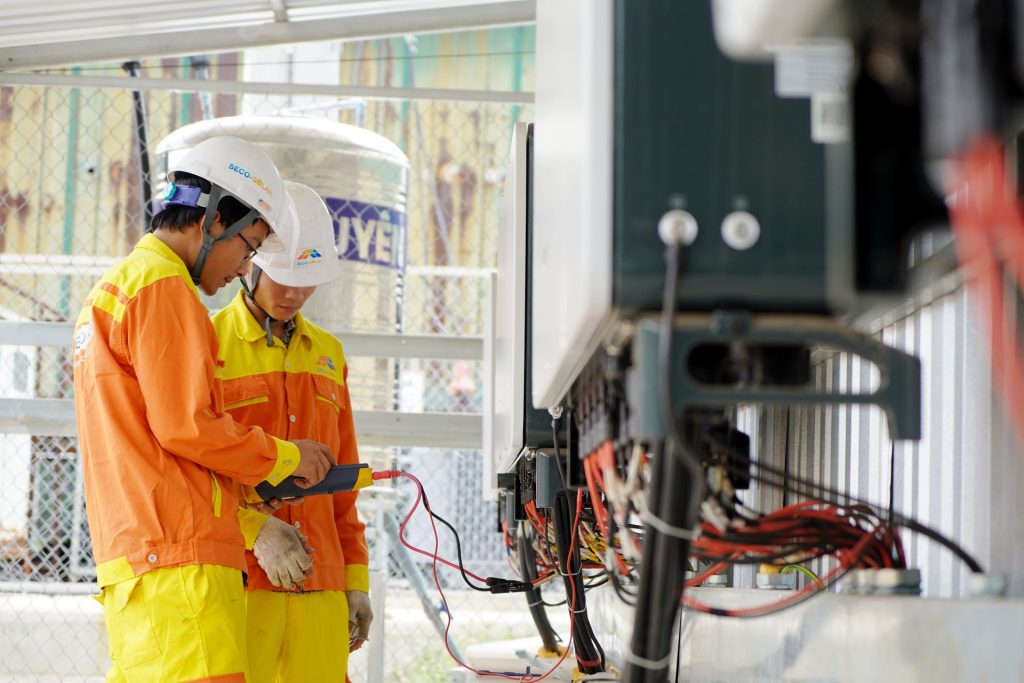 Two workers in orange and yellow safety gear and hard hats examine electrical equipment with tangled red and black cables at a construction site.